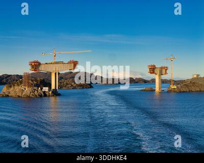 Küste, Norwegen - 25. November 2025: Eine Betonbrücke erhebt sich von felsigen Inseln mit Kranichen über einen Fjord an der norwegischen Küste, unter klarem blauem Himmel und klarem Winterlicht. Stockfoto