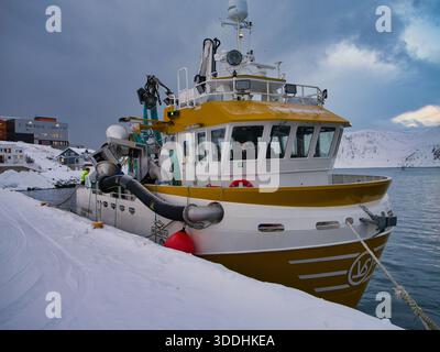 Honningsvag, Norwegen - 19. November 2025: Ein Fischerboot liegt an einem schneebedeckten Kai in Honningsvag, Norwegen, unter dramatischen Winterwolken mit arktischen Bergen in der Ferne Stockfoto