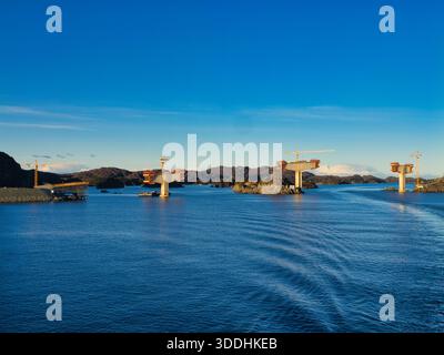 Küste, Norwegen - 25. November 2025: Brückenpfeiler und Turmkräne erheben sich aus einem Fjord an der Norways-Küste, die in klarem Winterlicht mit ruhigem blauem Wasser und zerklüfteten Inseln eingefangen werden. Stockfoto