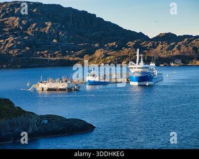 Küste, Norwegen - 25. November 2025: Dienstboote und schwimmende Käfige in einer Lachsfischfarm an der felsigen Küste Norwegens, beleuchtet durch klares Winterlicht und ruhiges blaues Wasser. Stockfoto