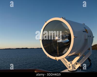 Küste, Norwegen - 25. November 2025: Großer Meeresscheinwerfer auf einem Schiffsdeck mit Blick auf die Küste Norwegens, beleuchtet von warmem, niedrigem Sonnenlicht vor einem klaren blauen Himmel. Stockfoto