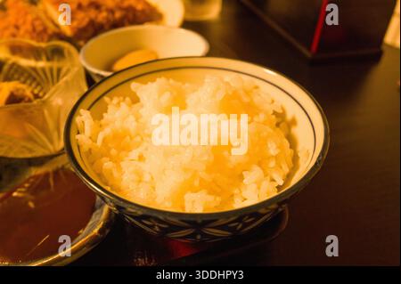 Schüssel mit flauschigem gedämpftem weißem Reis, serviert auf einem lackierten Tablett, umgeben von verschiedenen japanischen Beilagen unter warmer Beleuchtung in Hongu, Japan. Das Bild zeigt die Textur und Einfachheit der traditionellen japanischen Küche. Stockfoto