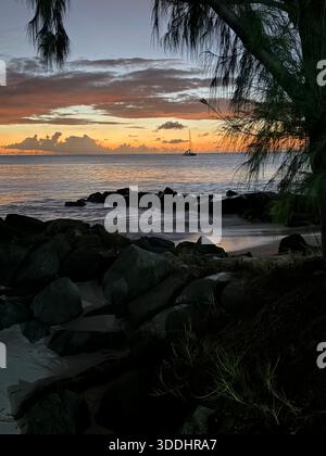 Segelyacht vor der Küste bei Sonnenuntergang über dunklen Felsen und überhängenden Küsten in Antigua, Karibik Stockfoto