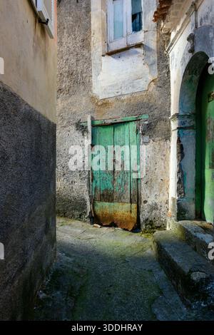 Verwitterte Holztür mit abblätternder grüner Farbe in einer rauen Steinfassade, beleuchtet durch nachmittags Sonnenlicht in einer schmalen Maratea Passage. Risse im Fenster und moosige Stufen verleihen dem Land einen rustikalen mediterranen Charakter. Stockfoto