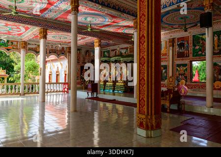 Geräumiger Saal eines buddhistischen Tempels in Vientiane, Laos, mit lebendigen erzählenden Wandgemälden, komplizierten Goldmustern und farbenfrohen Deckendesigns. Tageslicht unterstreicht den polierten Fliesenboden, die weißen Säulen und die kunstvollen Statuen und schafft eine ruhige und dekorative Atmosphäre. Stockfoto