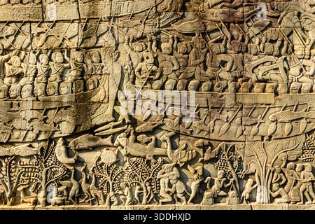 Detailliertes Sandstein-Basrelief zeigt eine alte Seeschlacht mit Soldaten, Booten, Fischen und Dschungeltieren an der äußeren Galeriemauer des Bayon-Tempels in Angkor, Kambodscha. Warmes Sonnenlicht unterstreicht die komplizierten Khmer-Schnitzereien und die strukturierte Oberfläche. Stockfoto