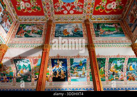 Lebendige erzählerische Wandmalereien und kunstvolle Muster schmücken die Innenwände und die Decke eines buddhistischen Tempels in Vientiane, Laos. Das komplexe Kunstwerk zeigt mythologische Szenen, Gottheiten und traditionelle Motive, die durch natürliches Licht und Goldakzente hervorgehoben werden. Stockfoto