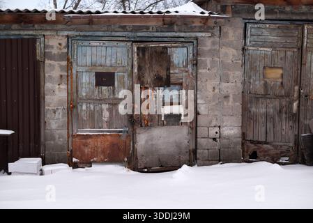 Verwitterte hölzerne Garagentüren im Winter. Alte verlassene Scheune mit abblätternder Farbe und rostigen Metallflecken, die mit Schnee bedeckt sind. Stockfoto