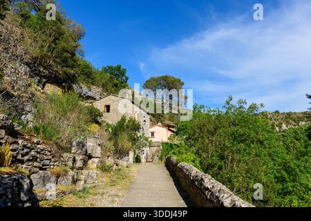 Ein schmaler Steinweg führt zu einem traditionellen Steinhaus, eingebettet zwischen felsigen Hügeln und üppigem Grün in Navacelles. Helles Sonnenlicht und klarer blauer Himmel unterstreichen die Texturen des Mauerwerks und der natürlichen Landschaft. Stockfoto