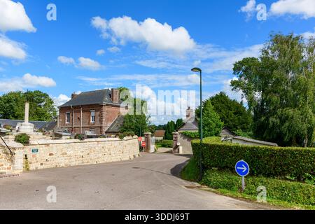 Eine friedliche ländliche Straße führt vorbei an einer Steinmauer und einem traditionellen Backsteinhaus, umgeben von üppigen Hecken und hohen Bäumen unter einem leuchtend blauen Himmel mit verstreuten Wolken. Die Szene bietet klares Sommerlicht und eine ruhige dörfliche Atmosphäre. Stockfoto