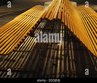 Gelbe Lattenbank, beleuchtet bei Nacht, wirft starke gestreifte Schatten über Pflastersteine in einem städtischen öffentlichen Raum. Stockfoto