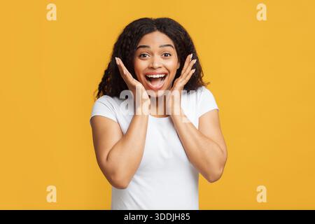 Wow-Angebot. Überglücklich afroamerikanische Dame, die die Kamera in Aufregung und berührendes Gesicht, gelber Studiohintergrund betrachte Stockfoto