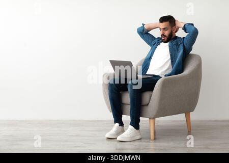 Schockierter junger arabischer Mann, der seinen Kopf in der Nähe eines Laptops schnappte und Fehler bei einem Online-Job-Projekt in der Nähe einer weißen Studiowand machte Stockfoto