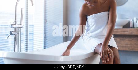 Frau in weißem Handtuch sitzt tagsüber neben einer freistehenden Badewanne in einem modernen Badezimmer Stockfoto