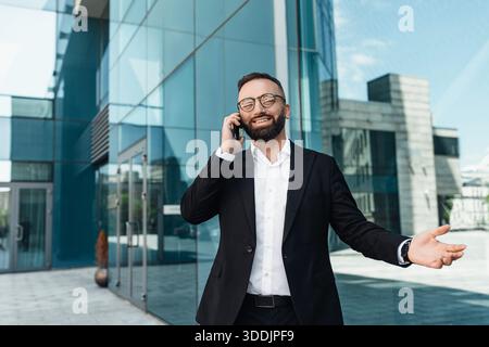Geschäftsgespräch. Glücklicher, reifer Unternehmer, der auf dem Smartphone spricht und im Freien gegen das Bürozentrum steht Stockfoto
