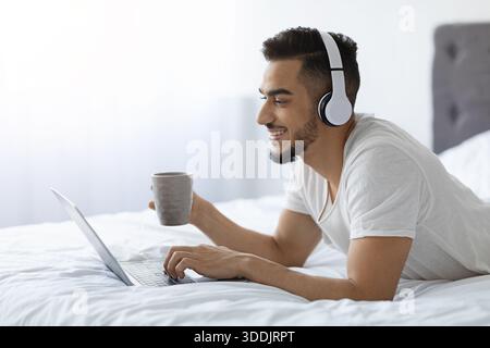 Millennial arabischer Mann, der Kaffee genießt, während er Laptop im Bett benutzt Stockfoto