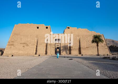 Eine weite Aufnahme, die eine Person in traditionellen blauen Gewändern zeigt, die in Richtung des massiven Eingangs des Grabentempels von Ramses III. Geht Stockfoto