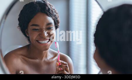 Frau putzt Zähne im Badezimmerspiegel während der Morgenroutine mit Zahnspangen Stockfoto