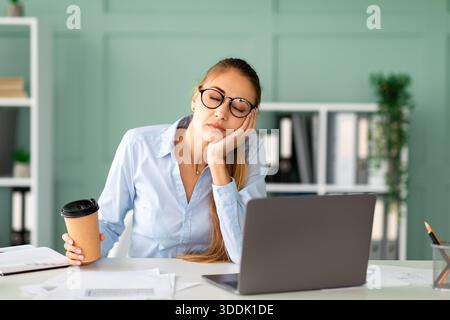 Junge Frau im Büro, die nach langer Computerarbeit schläft, während sie Kaffeetasse hält Stockfoto