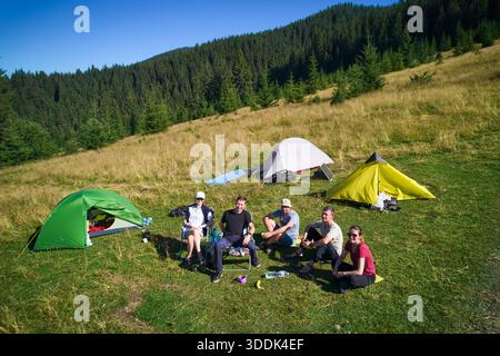 Campingplatz aus der Vogelperspektive in den Bergen, mit mehreren bunten Zelten auf einer grasbewachsenen Rodung. Camper entspannen unter klarem blauen Himmel, umgeben von hohen immergrünen und Panoramablick auf entfernte Gipfel. Stockfoto
