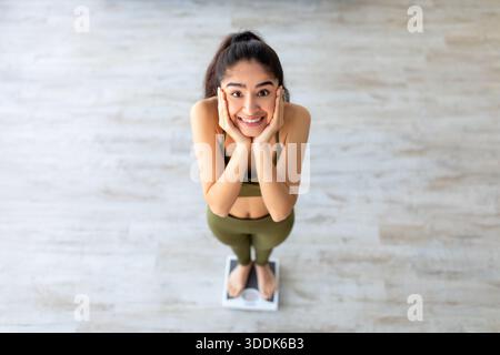 Hübsche indische Frau auf einer Waage, die ihr Gesicht in Aufregung hält und nach erfolgreichem Abnehmen zu Hause, über dem Bild, wiegt Stockfoto