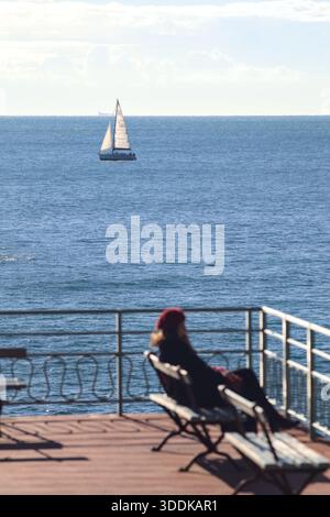 Segelboot auf dem Meer, von einem Balkon aus gesehen, mit einer unfokussierten Frau, die auf einer Bank sitzt Stockfoto