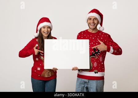 Glückliches Paar mit weihnachtlichen Pullover und Hüten, die ein leeres Schild für festliche Ankündigungen halten Stockfoto