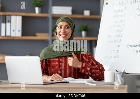 Islamischer Lehrer im Kopftuch, der am Schreibtisch sitzt und den Daumen nach oben zeigt Stockfoto