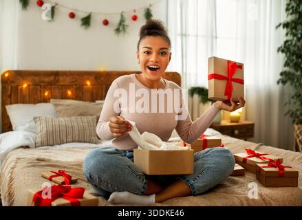 Portrait einer schwarzen Dame, die Weihnachtsgeschenke erhält und Pappkarton auspackert, die im gemütlichen Schlafzimmer auf dem Bett sitzt Stockfoto