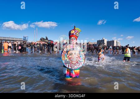 Eine Person in einem Clownkostüm mit einer großen kreisförmigen Requisite mit Gesicht und Text steht im Meer vor einer Menschenmenge. Stockfoto