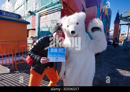 Ein Mann in orangefarbenen Hosen und einer schwarzen Jacke hält ein „DO IT!“ Zertifikat neben einer Person in einem weißen Eisbärenkostüm. Stockfoto
