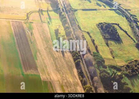 Blick aus der Vogelperspektive auf ländliches Ackerland zur goldenen Stunde, mit Flickenteppichfeldern aus lebhaftem Grün und brauner Erde. Kurvenreiche Autobahn durchquert die Linie Stockfoto