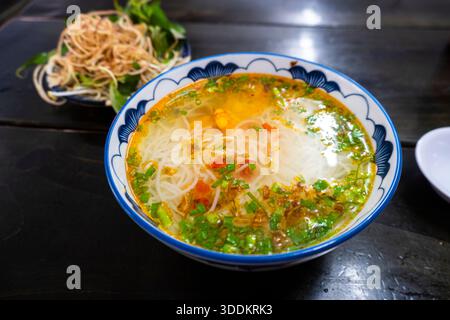 Eine Schüssel Suppe mit Nudeln und grünen Kräutern steht auf einem Tisch. Die Schüssel ist blau und weiß, und die Suppe ist klar Stockfoto
