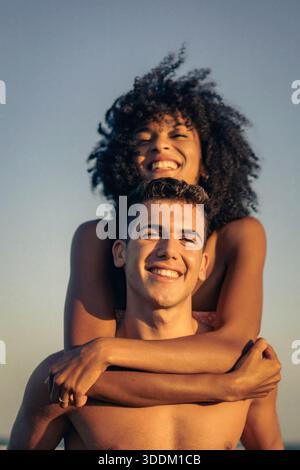 Ein fröhliches, abwechslungsreiches Paar teilt sich einen Huckepack-Ritt im Freien und lacht während der goldenen Stunde glücklich. Authentisches Lächeln fängt einen Moment puren Spaßes und c ein Stockfoto