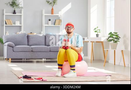 Lustiger verärgerter Mann in Sportbekleidung, der auf passendem Ball sitzt und Hanteln hält, müde vom harten Workout zu Hause Stockfoto
