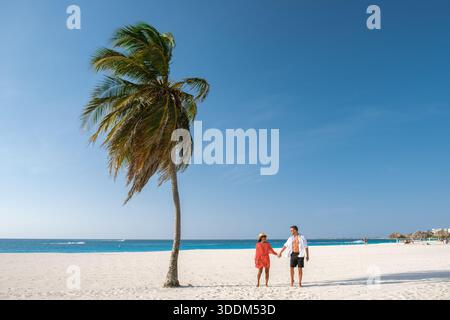 An einem sonnigen Tag am Eagle Beach macht ein Paar einen romantischen Spaziergang, Hand in Hand, unter dem sanften Schaukeln der Palmen. Der goldene Sand und das klare blaue Wasser schaffen einen perfekten tropischen Zufluchtsort in Aruba. Stockfoto