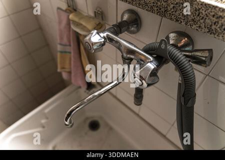 Chrom-Badezimmerarmatur mit Kunststoffschlauch und Duschkopf, der an einer gefliesten Wand über einem Keramikwaschbecken befestigt ist und alte Sanitär- und Renovierungsarbeiten darstellt Stockfoto