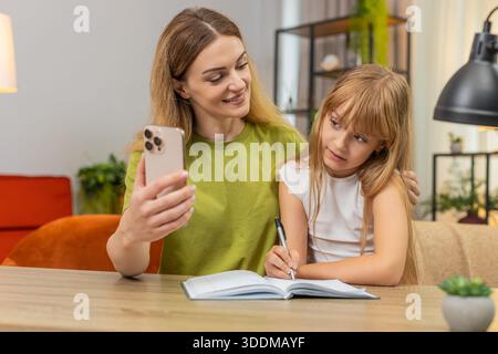 Mutter und Tochter sehen zu Hause Online-Unterricht auf dem Smartphone Schreiben Sie Notizen in Notizbuch Folgen Sie Aufgaben. Frau und Mädchen am Tisch überprüfen Übung markieren Antworten Fehler beheben Regeln Praxis Fokus Tempo gut lernen Stockfoto