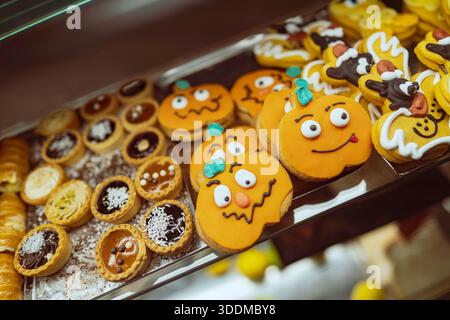 Verschiedene Kürbiskekse mit halloween-Thema und lustigen Gesichtern und verschiedenen süßen Torten füllen ein klares Regal und präsentieren festliche Backwaren für einen Stockfoto