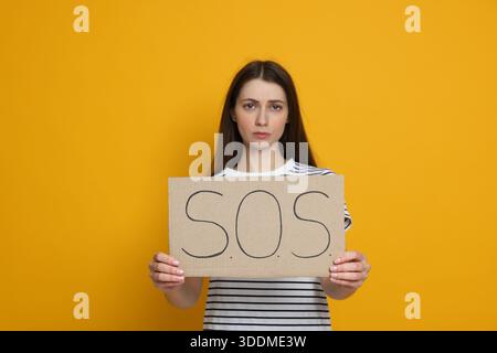 Besorgte Frau, die ein Schild mit dem Wort SOS auf gelbem Hintergrund hält Stockfoto