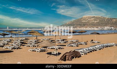 Eine friedliche Küstenszene mit einem roten Fußweg, der zum sonnendurchfluteten Horizont führt, Fahnen wehen, gestreiften Sonnenschirmen und leeren Stühlen, die auf Freizeit hinweisen, trav Stockfoto