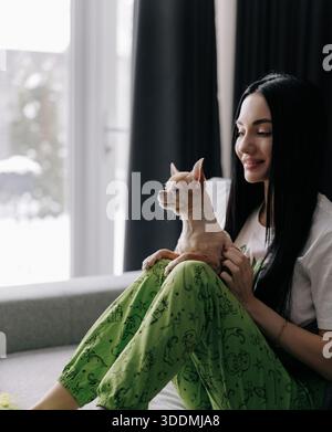 Seitenansicht einer lächelnden Frau mit langen dunklen Haaren, die sich auf der grauen Couch entspannt und einen kleinen, braunen chihuahua auf ihrem Schoß hält. Frau trägt grünen Pyjama in Mo Stockfoto