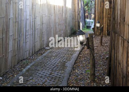 Vintage Lampenpfosten auf Bambuszaun Gehweg mit ruhiger Gartenatmosphäre in der Nähe von perforiertem Betonblockweg mit geheimnisvollem Abendlicht und rustikalem Stall Stockfoto