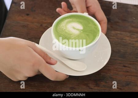 Entspannende Hände halten warmen, grünen Matcha-Tee-Latte mit Milchschaumkunst in weißer Keramikuntertasse auf Holztisch und sorgen für ein gemütliches, komfortables Gefühl Stockfoto