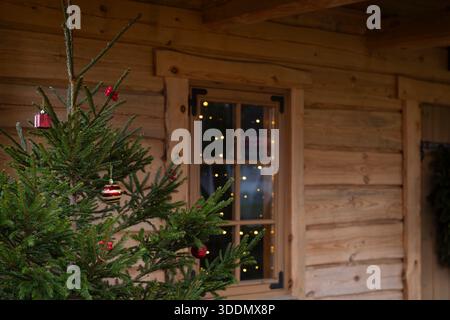 Weihnachtsbaum steht vor einer Holzhütte, mit warmen festlichen Lichtern, die durch das Fenster leuchten. Gemütliche rustikale Hütte außen mit einem Weihnachtsbaum. Stockfoto