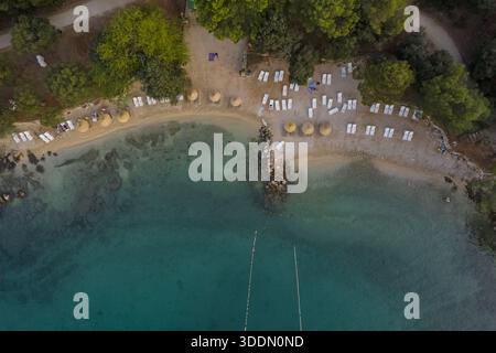 Blick aus der Vogelperspektive auf den sonnenverwöhnten Strand mit Reihen von weißen Liegestühlen und Strohschirmen, die auf das türkisfarbene Wasser treffen, Rovinj, Kreis Istrien, Kroatien. Stockfoto