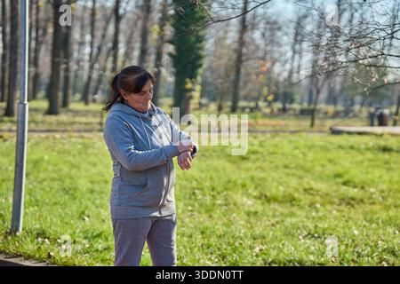 Eine ältere Frau überprüft ihre Smartwatch-Fitness-Statistiken nach einem Lauf im Park an einem sonnigen Morgen Stockfoto