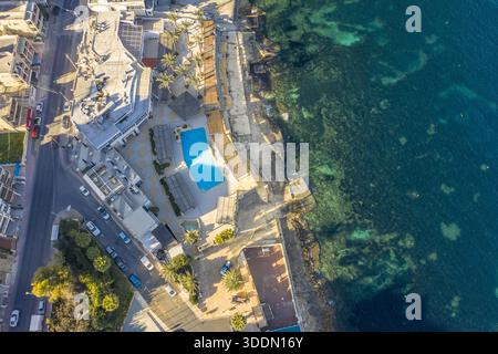 Aus der Vogelperspektive auf türkisfarbenes Wasser trifft auf die zerklüftete Küste in der Nähe eines Hotels mit einem hellblauen Pool, Bugibba, St. Paul's Bay, Malta. Stockfoto