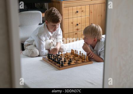 Zwei Brüder sitzen auf dem Bett und spielen zu Hause Schach. Sie konzentrieren sich auf Bord mit nachdenklichen Ausdrücken bei weichem Tageslicht Stockfoto