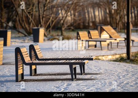 Leere hölzerne Liegen zum Ausruhen stehen in der Nähe des Flusses im Stadtpark bedeckt mit Schnee an sonnigen Wintertagen. Stockfoto